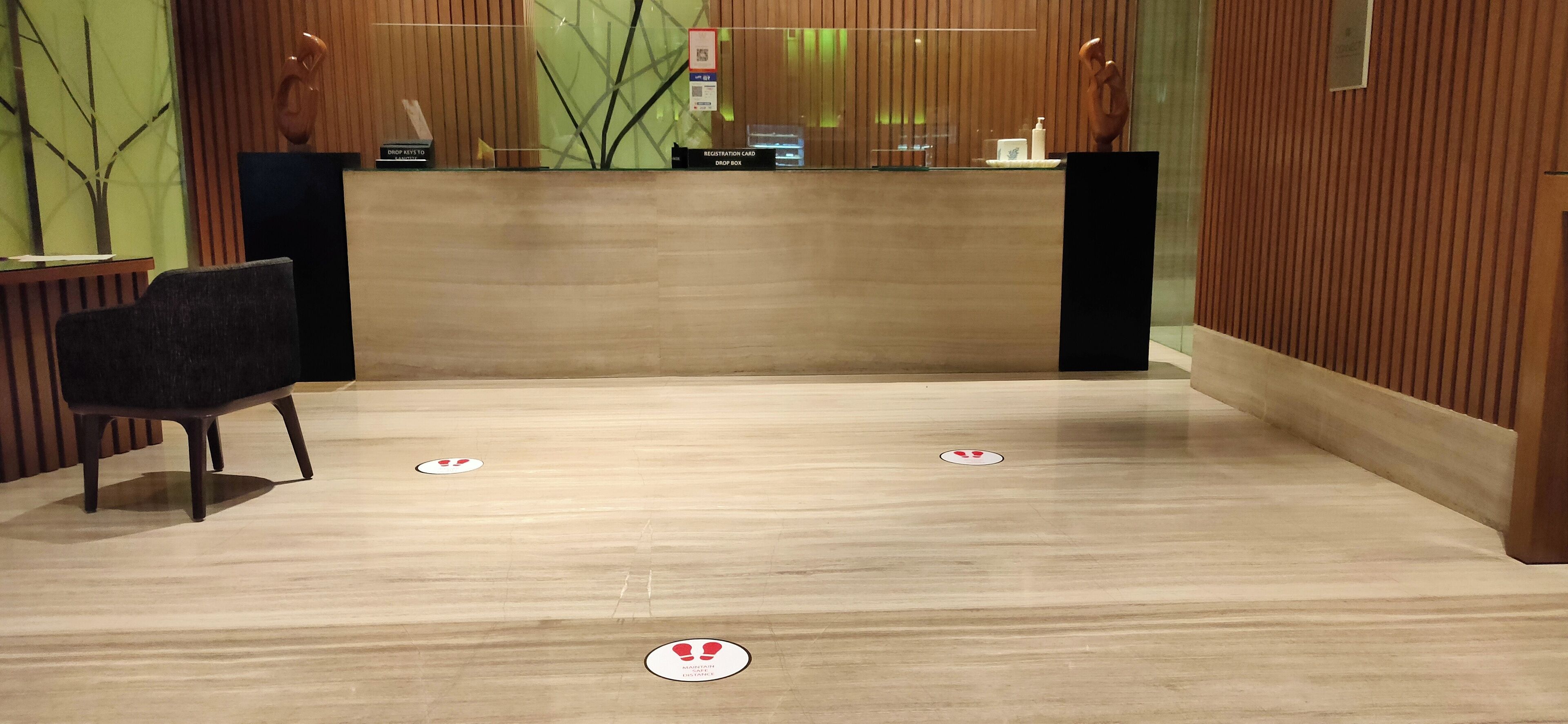 A reception area with a wooden reception desk, a black chair, and a glass partition. There are signs on the floor indicating social distancing measures.