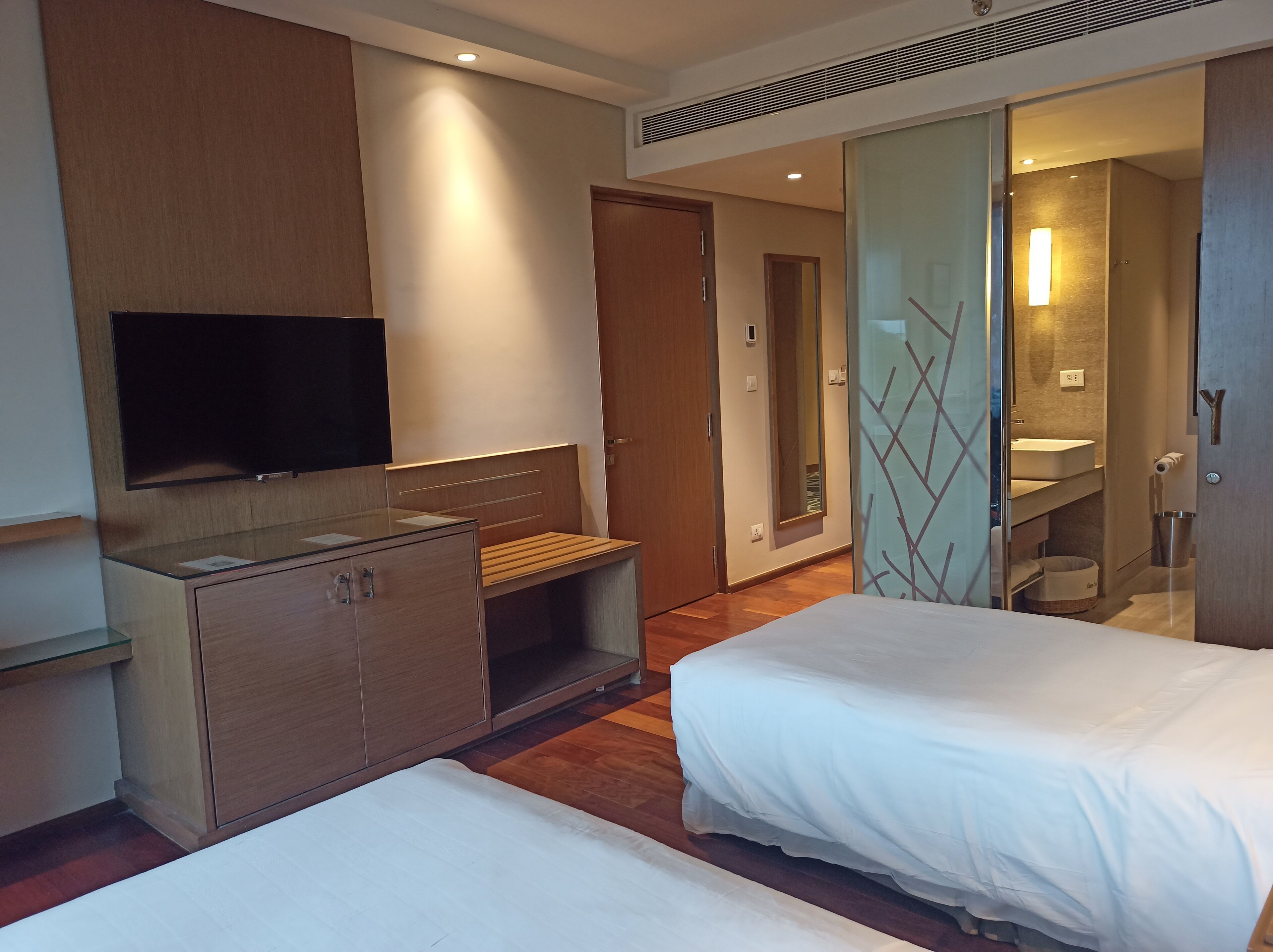 A hotel room with a bed, a TV, a desk, and a bathroom visible through a glass door.
