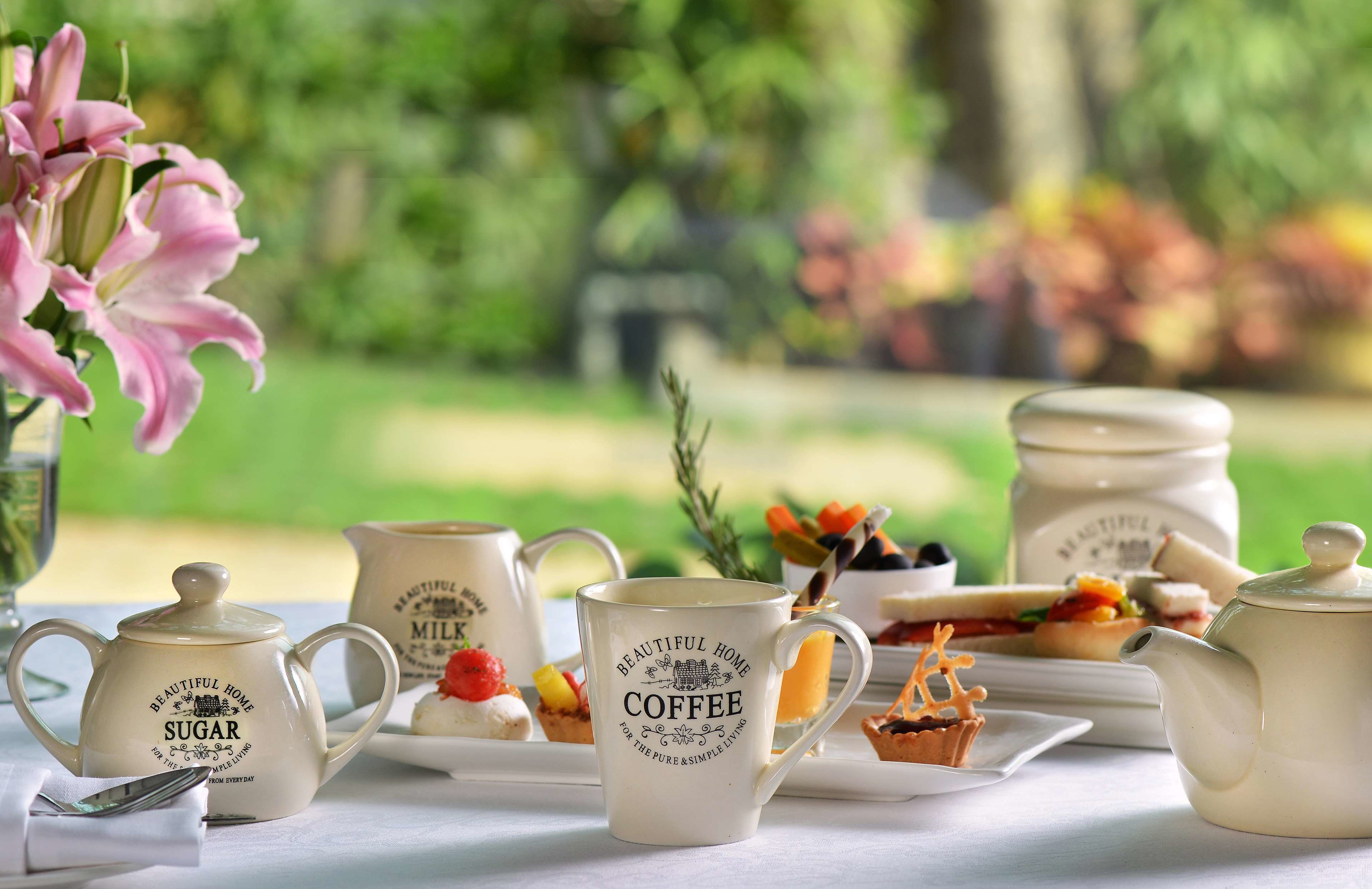 A tea set with sugar and coffee mugs, a teapot, and a sugar bowl on a table with a floral arrangement and a view of a garden.