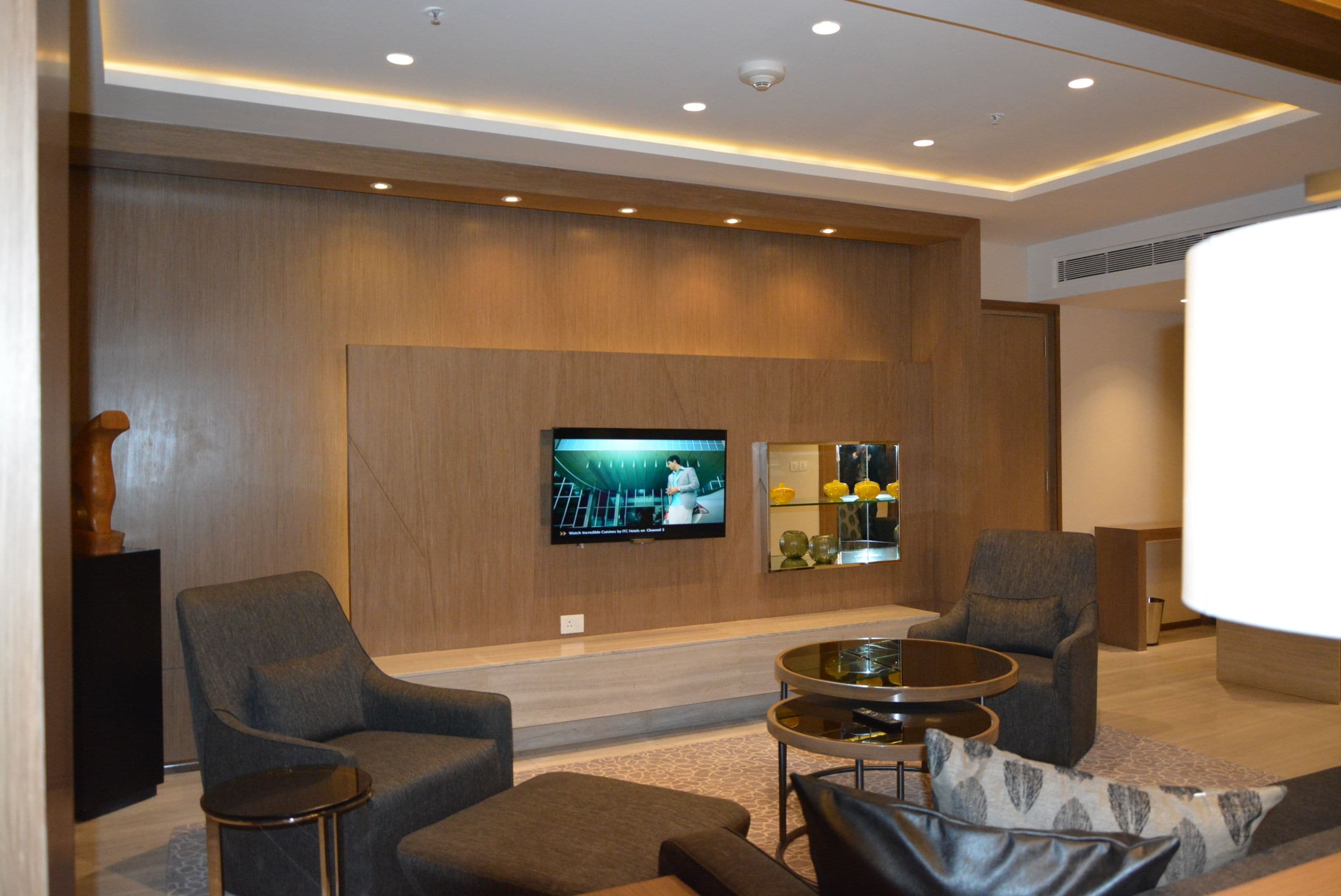 A modern hotel lobby with a flat-screen TV mounted on the wall, two armchairs, a small round table, and a sofa.