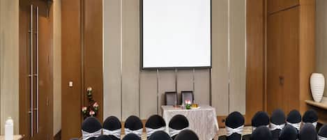 Un salón de conferencias con filas de sillas, una pantalla de proyector y una mesa con un arreglo floral.