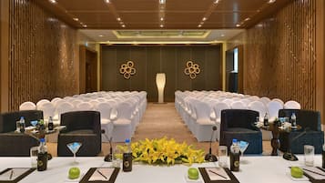 Un salón de conferencias con filas de sillas blancas, un escenario con un atril y arreglos florales sobre las mesas.