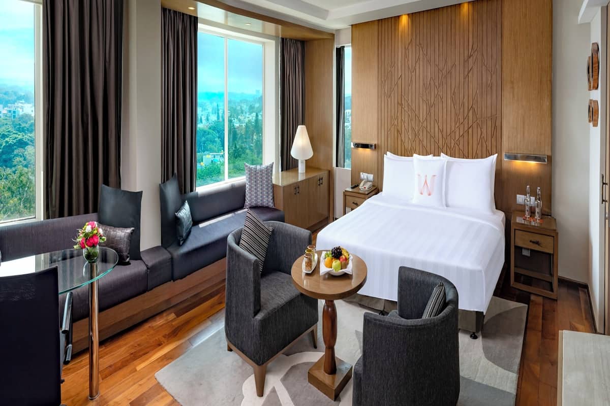 A modern hotel room with a large bed, a sofa, two armchairs, a glass table, and a view of the city.