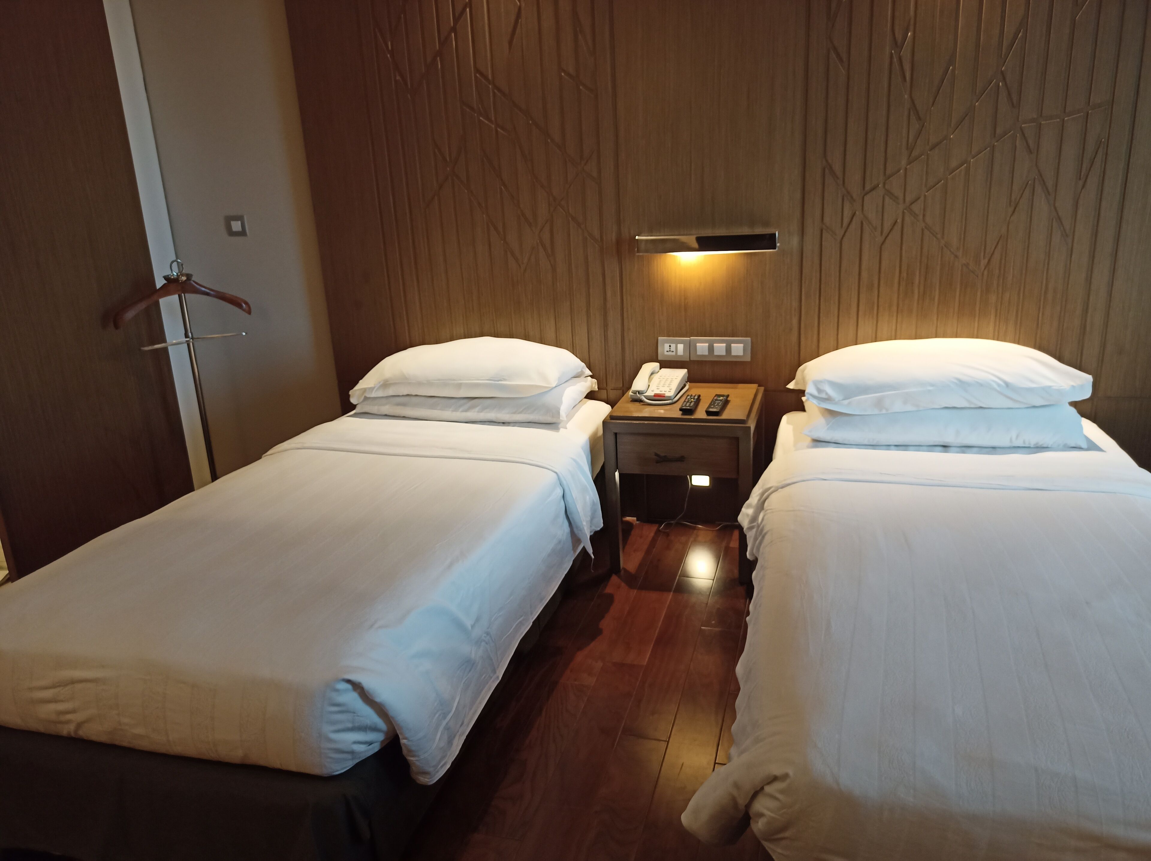 Une chambre d’hôtel avec deux lits simples, une armoire en bois, une table de chevet avec un téléphone et une télécommande, et un luminaire fixé au mur.