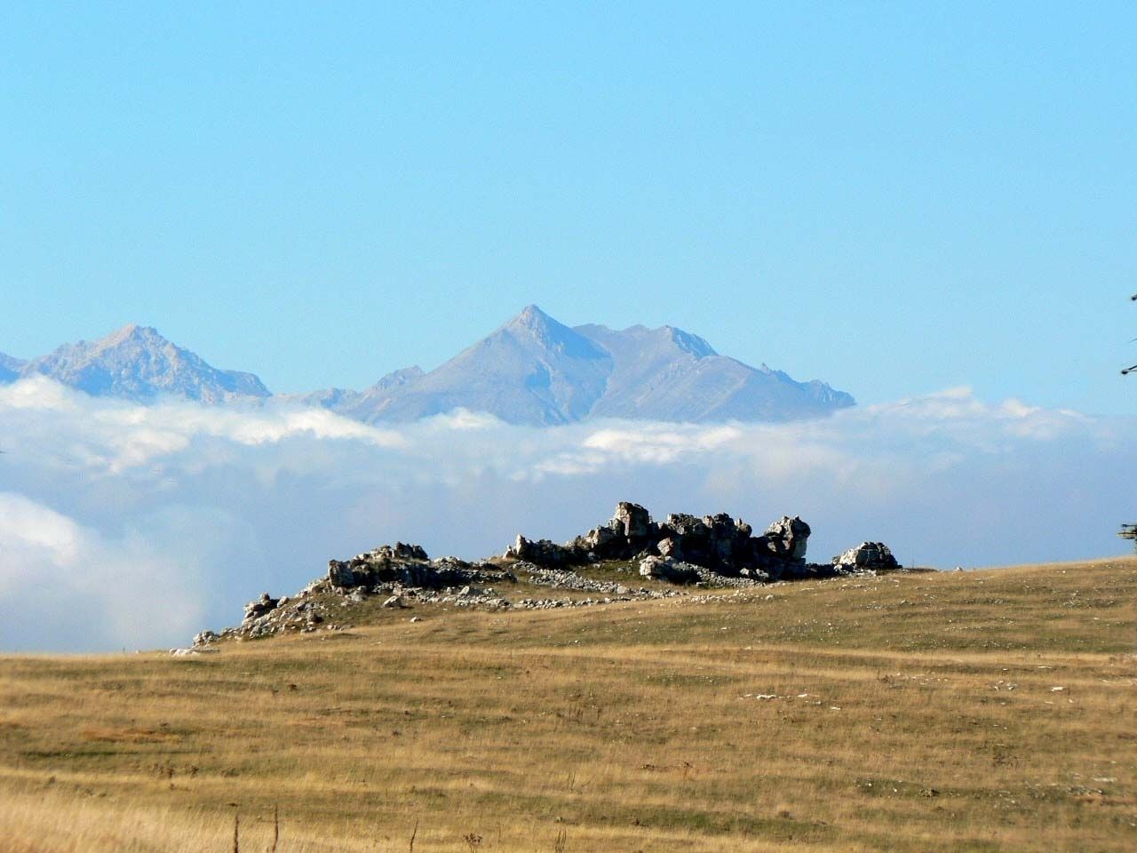 Un paisaje montañoso con un afloramiento rocoso en primer plano y cumbres nevadas al fondo.