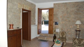 A stone-walled room with a wooden door, a radiator, a table with a vase of flowers, and a lamp.