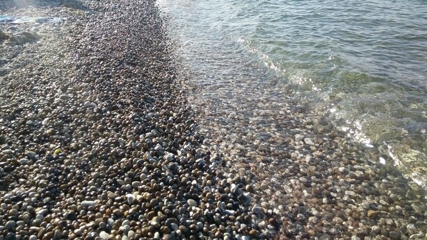 A pebbled beach with clear water, rocky shore, and distant hills.