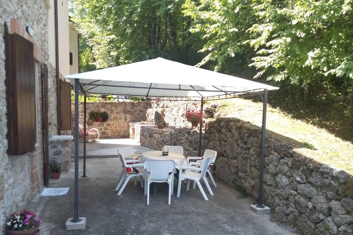 An outdoor patio with a table and chairs under a white canopy, surrounded by stone walls and greenery.