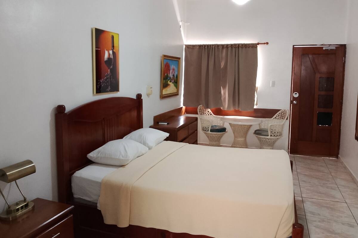 A hotel room with a bed, bedside table, lamp, and a small seating area with a table and chairs.