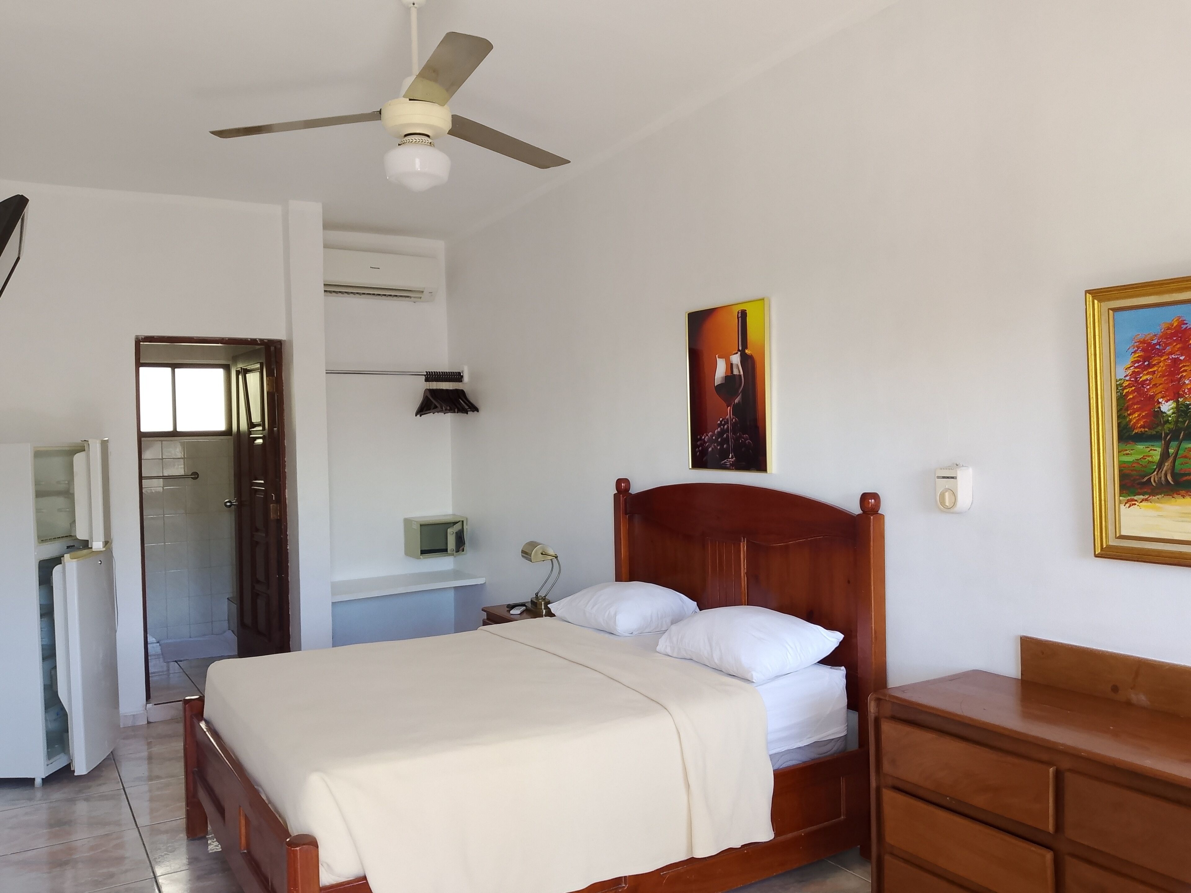 Ein ordentlich eingerichtetes Schlafzimmer mit Bett, Nachttisch und Kommode. Es gibt einen Deckenventilator, eine Wandklimaanlage und ein Bild an der Wand.