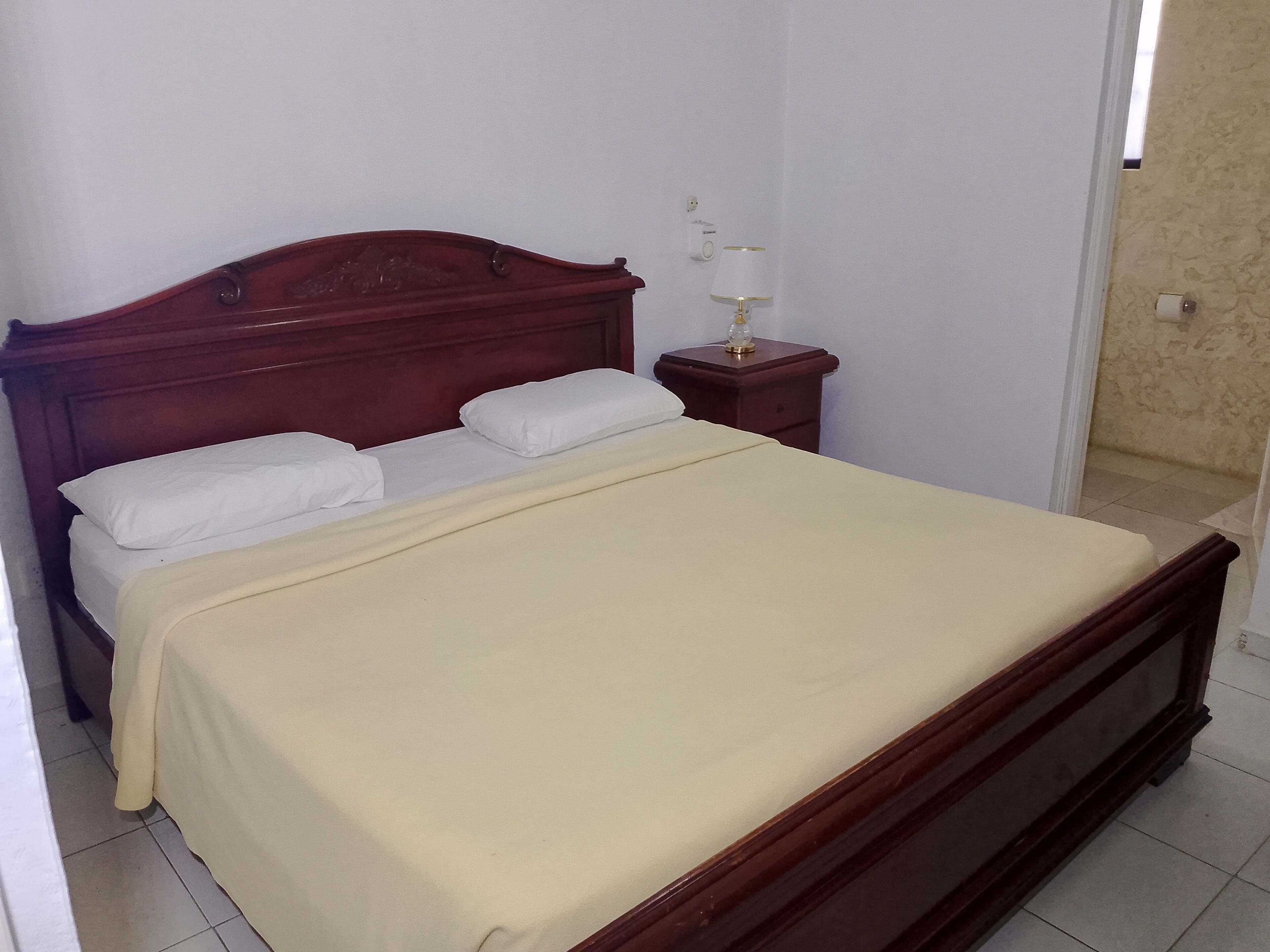 A single bed with a wooden headboard, white pillows, and a beige bedspread in a room with white walls and a tiled floor.