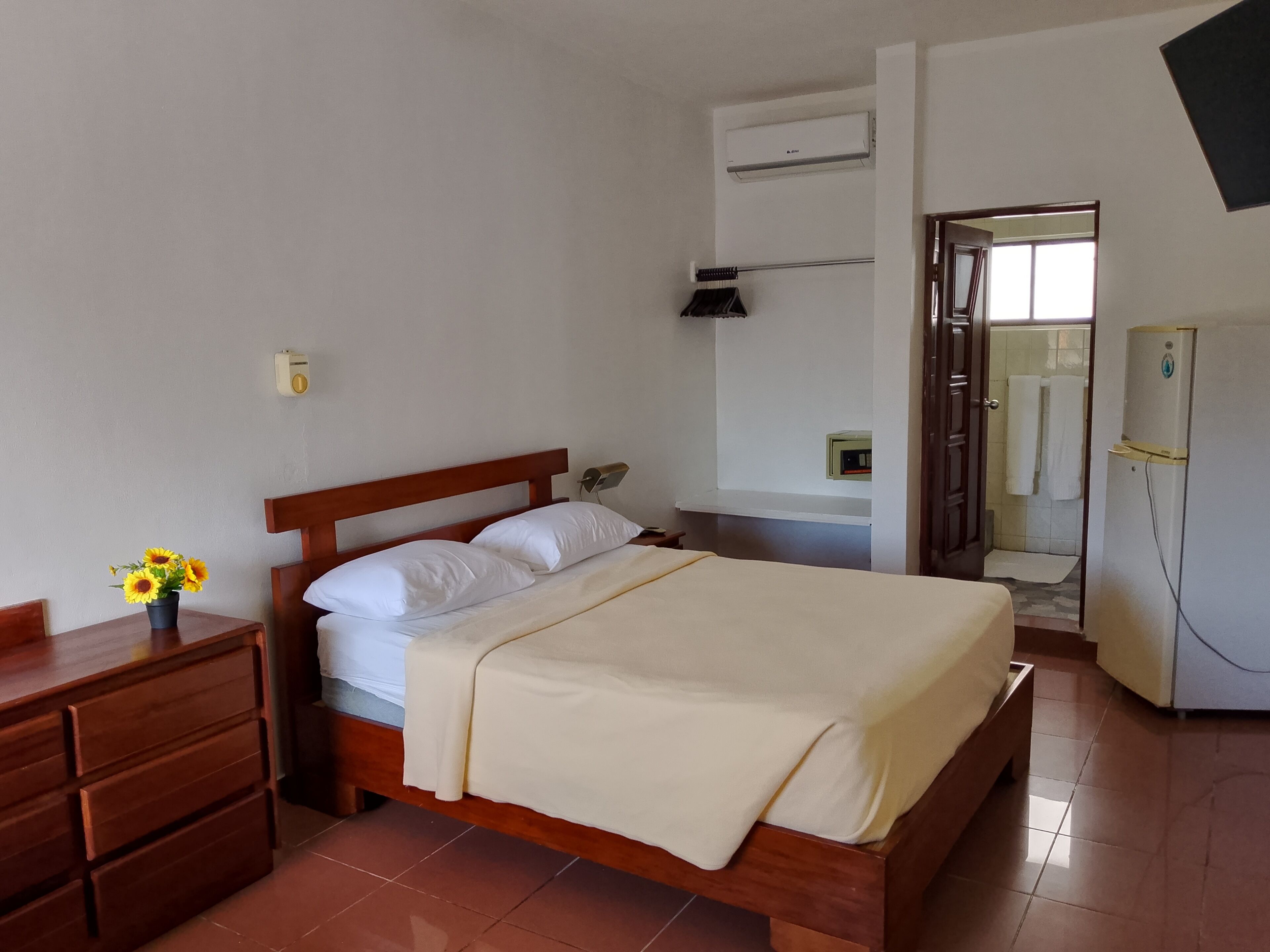 A bedroom with a wooden bed, white bedding, a nightstand with a vase of sunflowers, a wall-mounted air conditioner, and a bathroom visible through an open door.