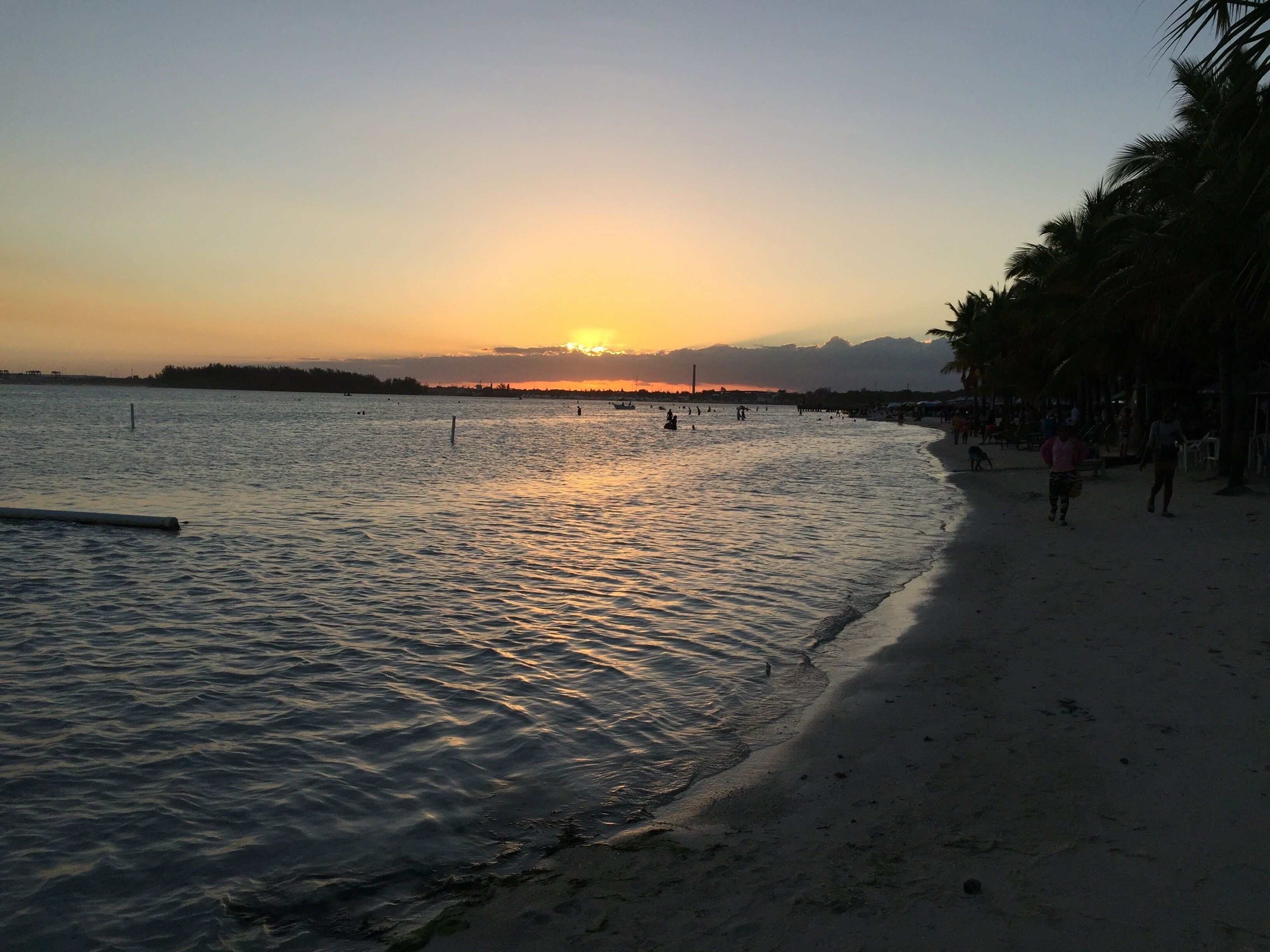 A beach with calm water, a sunset, and people walking along the shore.