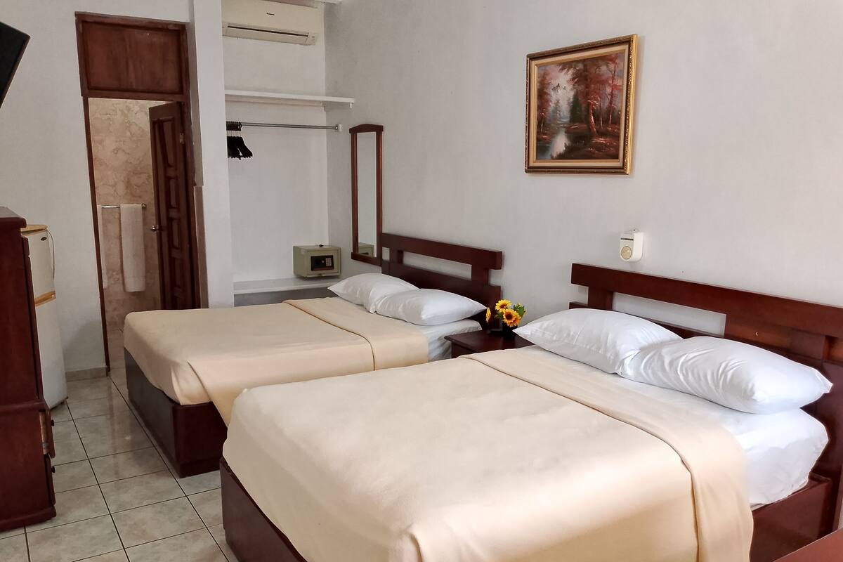 A hotel room with two beds, a wooden wardrobe, a mirror, a painting on the wall, and a small table with a vase of flowers.