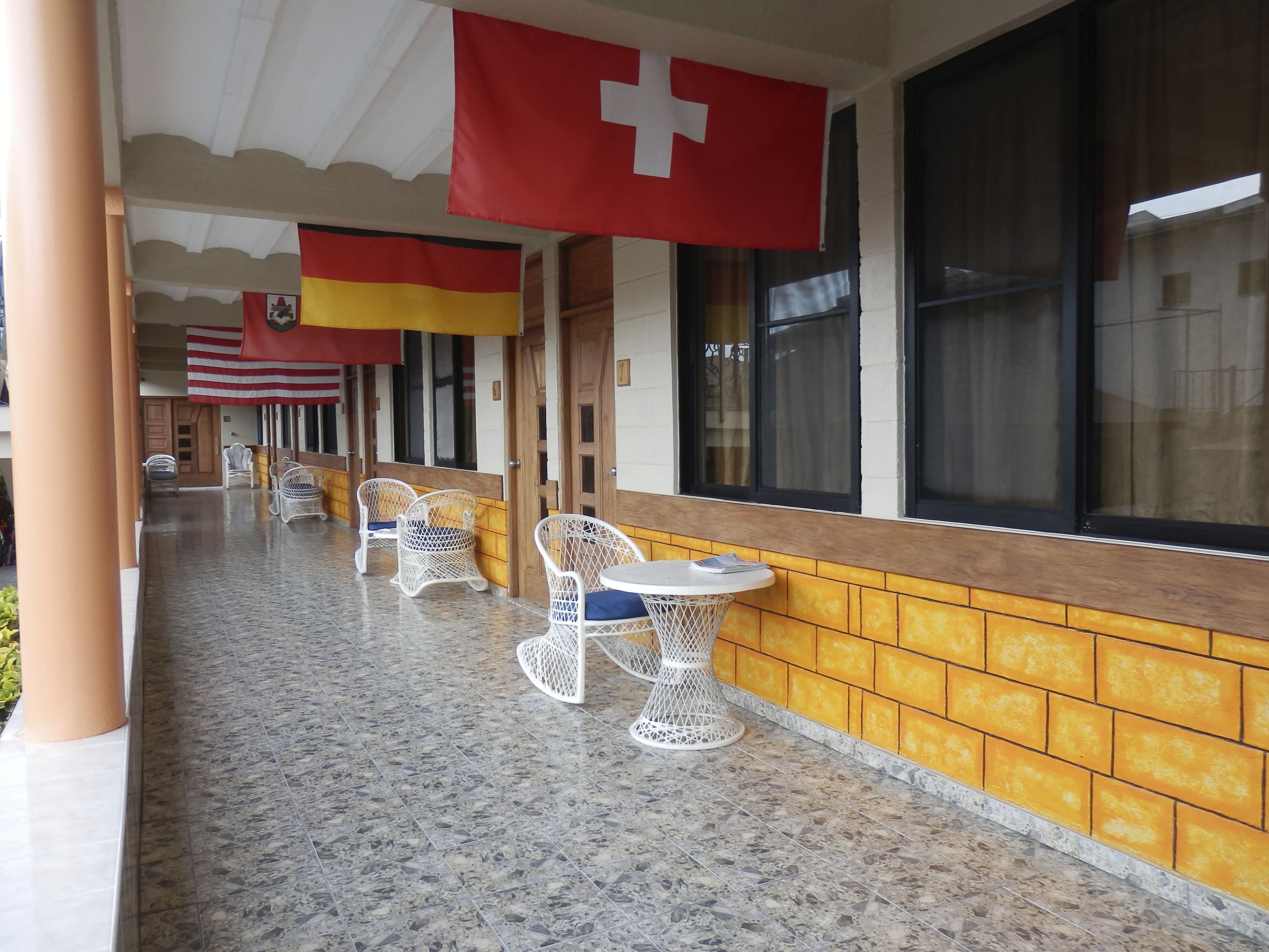 A covered outdoor seating area with wicker chairs and tables, a tiled floor, and large windows with curtains.