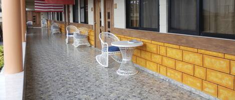 A covered outdoor seating area with wicker chairs and tables, a tiled floor, and large windows with curtains.