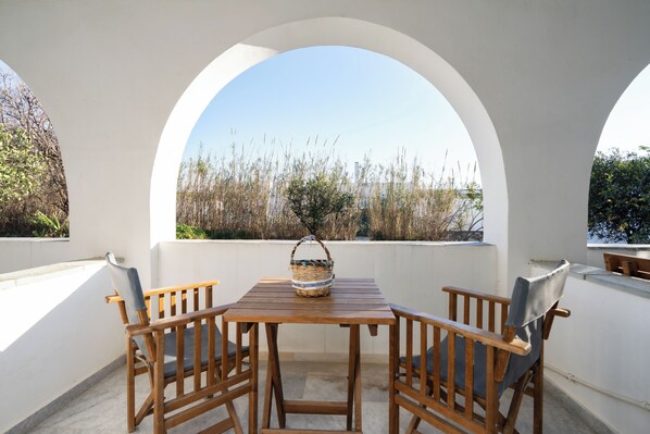 Una mesa de madera con sillas y una cesta en un balcón con arcos blancos y vistas a pastizales altos.