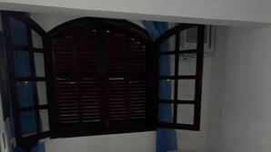 Una cama pequeña y bien arreglada con colcha a cuadros y dos almohadas, un rascador para gatos negro y una ventana con contraventanas de madera.