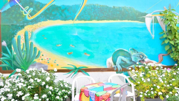 Un mural colorido muestra una escena de playa con una tienda de campaña, un paracaídas y un bote. Se ven sillas blancas y una mesa con un mantel de colores.
