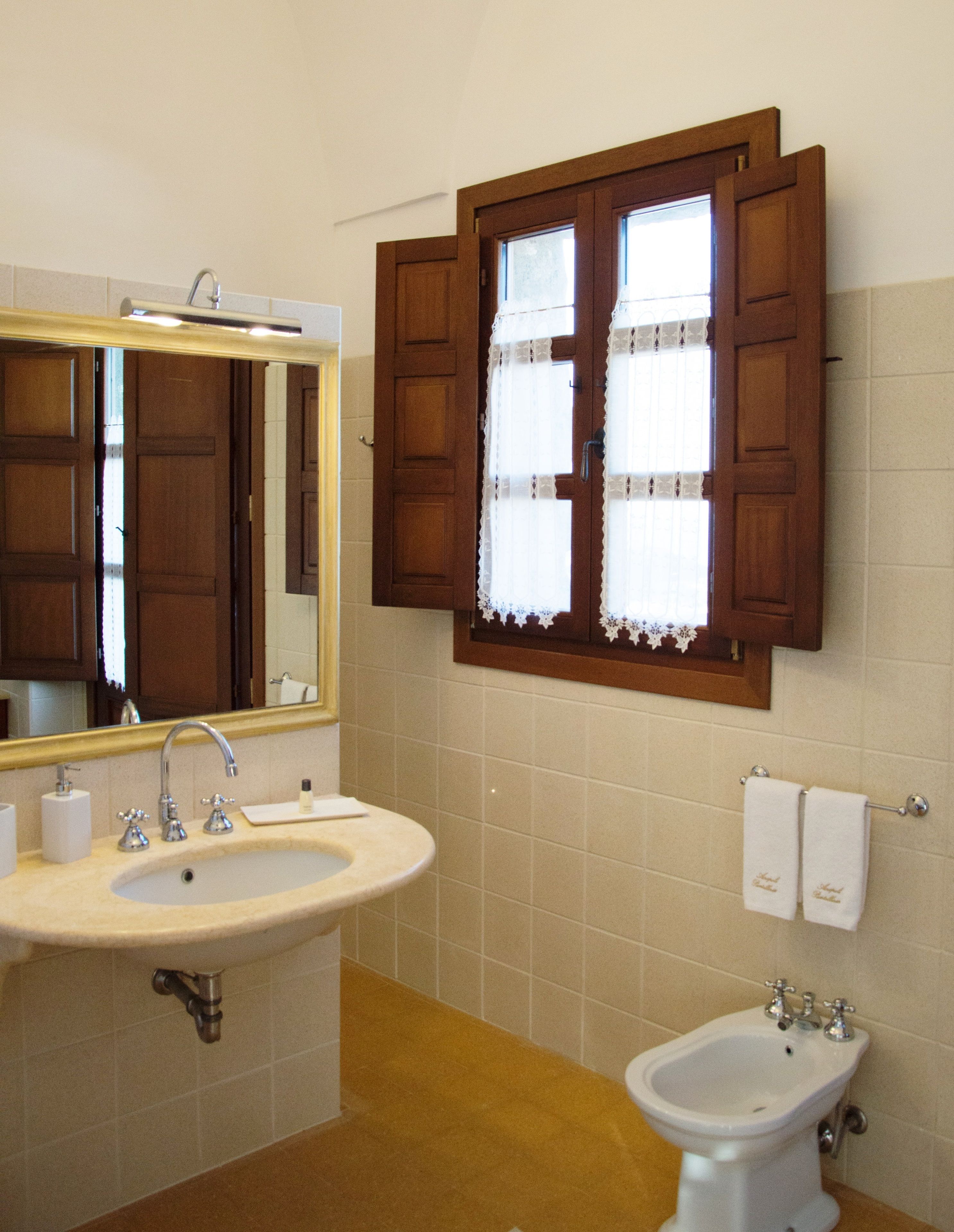 A bathroom with a sink, bidet, mirror, and wooden window shutters.