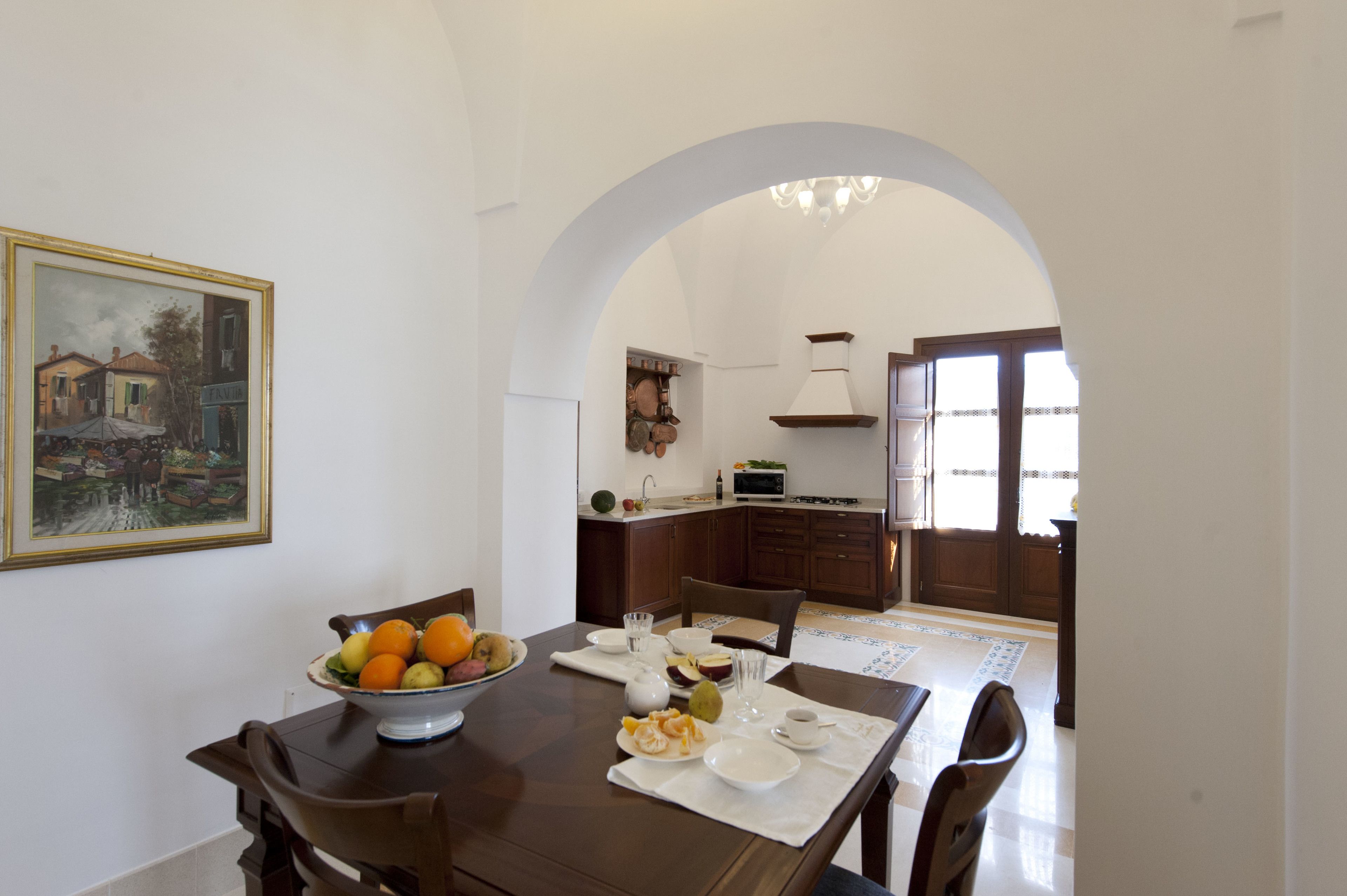 A dining area with a table set for two, a bowl of fruit, and a painting on the wall.