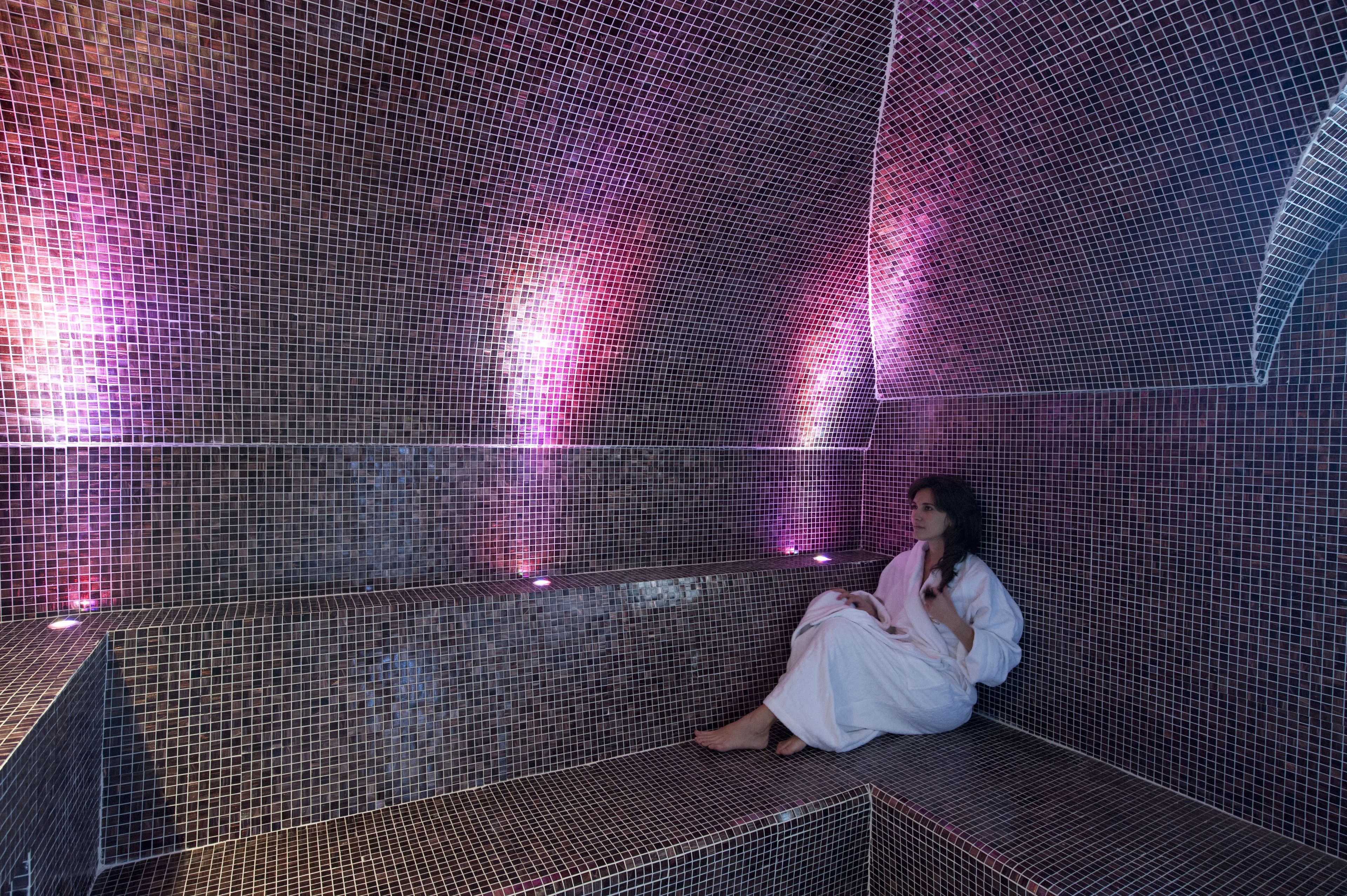 A person sitting in a tiled room with a curved ceiling and pink lighting.