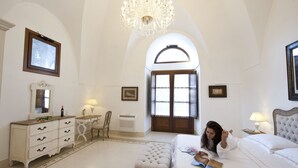 A spacious bedroom with a large bed, a vanity with a mirror, a chandelier, and a view through an arched window. - Resort Acropoli (Pantelleria)