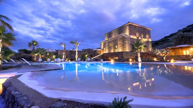 A large swimming pool with lounge chairs and palm trees, surrounded by stone buildings with warm lighting. - Resort Acropoli (Pantelleria)