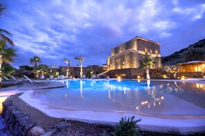 A large swimming pool with lounge chairs and palm trees, surrounded by stone buildings with warm lighting. - Resort Acropoli (Pantelleria)
