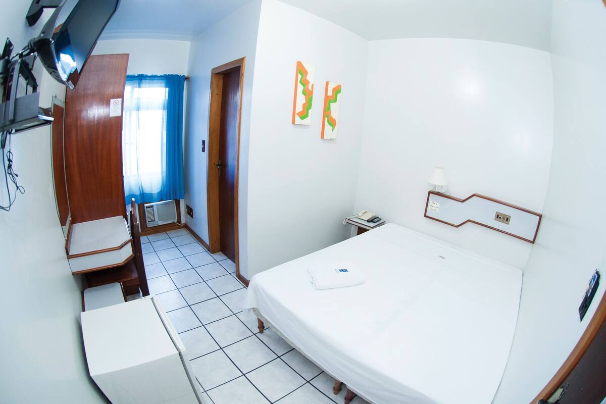 A hotel room with a bed, a desk, a chair, a small table, a lamp, and a window with curtains.