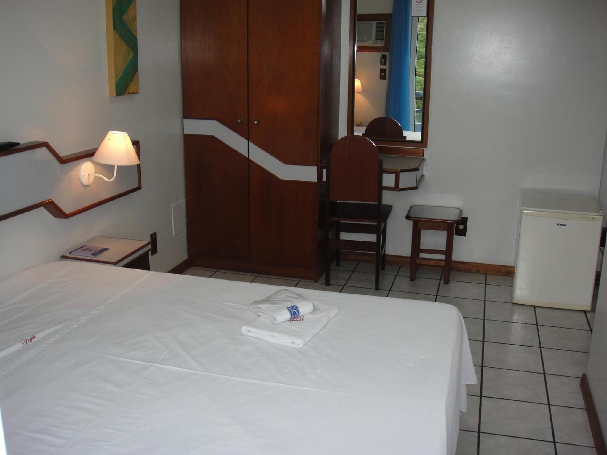 Habitación de hotel con cama, escritorio, silla, mesita, lámpara y refrigerador.