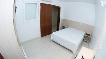 A hotel room with a bed, a wooden wardrobe, a small bedside table, a desk with a chair, and a window with blinds.