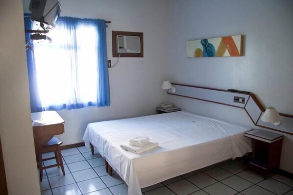 Habitación de hotel con cama, escritorio, silla, mesita de noche, lámpara y ventana con cortinas.