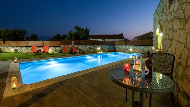 A swimming pool with a stone wall and wooden deck, surrounded by lounge chairs and a table with drinks. - Skandalis Traditional Suites (Chania)