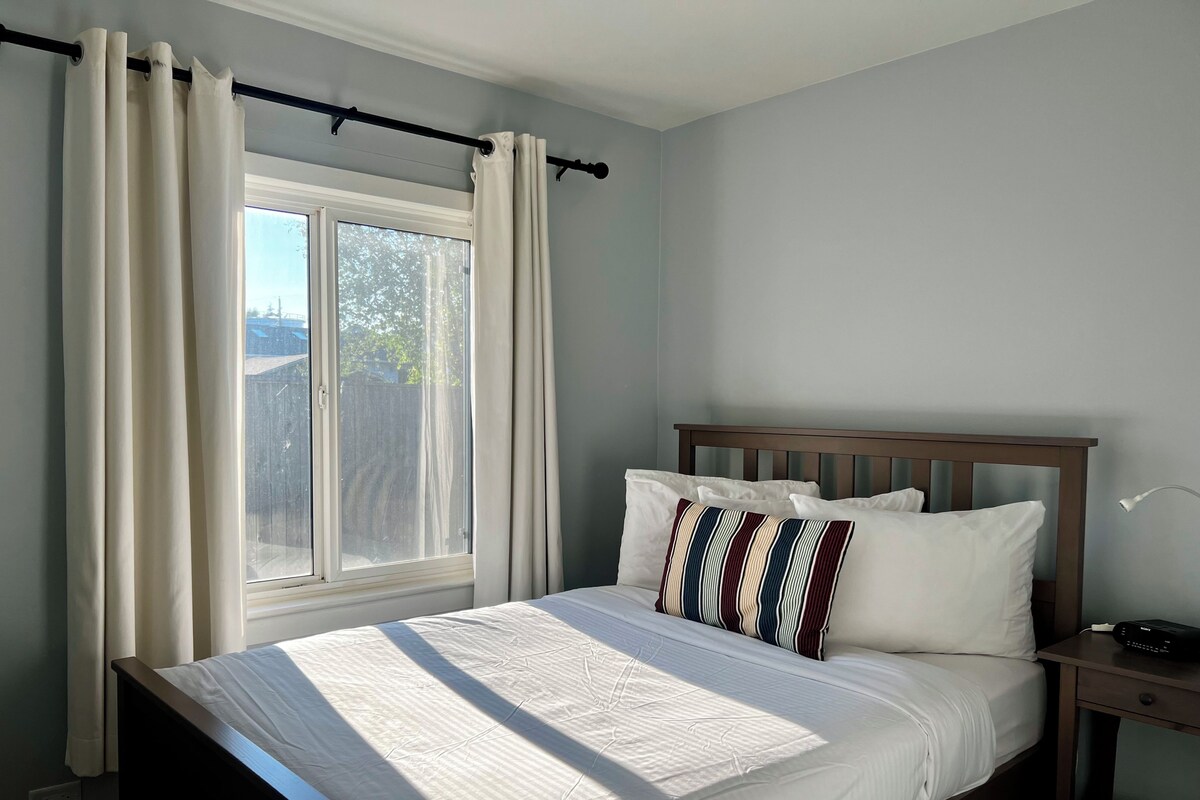 Une chambre à coucher avec un lit, une fenêtre avec des rideaux, une table de chevet avec une lampe, et une vue sur l’extérieur.