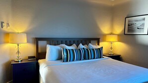 A neatly made bed with white linens and striped pillows, flanked by two bedside tables with lamps, in a room with a textured wall and a framed picture above the bed. - Buccaneer Inn (Nanaimo)