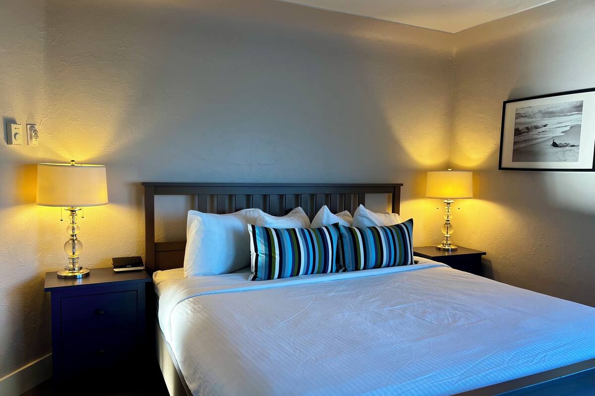 A neatly made bed with white linens and striped pillows, flanked by two bedside tables with lamps, in a room with a textured wall and a framed picture above the bed.