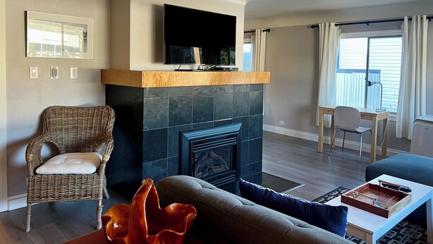 A living room with a fireplace, a television mounted above it, a wicker chair, a sofa, and a dining area with a table and chairs. - Buccaneer Inn (Nanaimo)