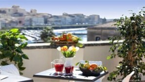 A table set with tea cups, a teapot, and a fruit basket on a balcony with a city view. - Hotel Le Muse (Syracuse)