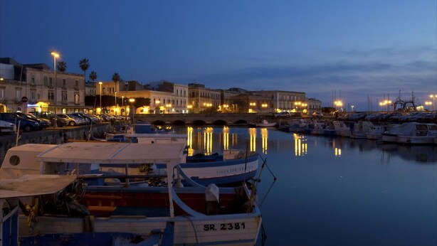 A marina at dusk with boats docked, a calm water surface reflecting lights, and buildings in the background. - Hotel Le Muse (Syracuse)