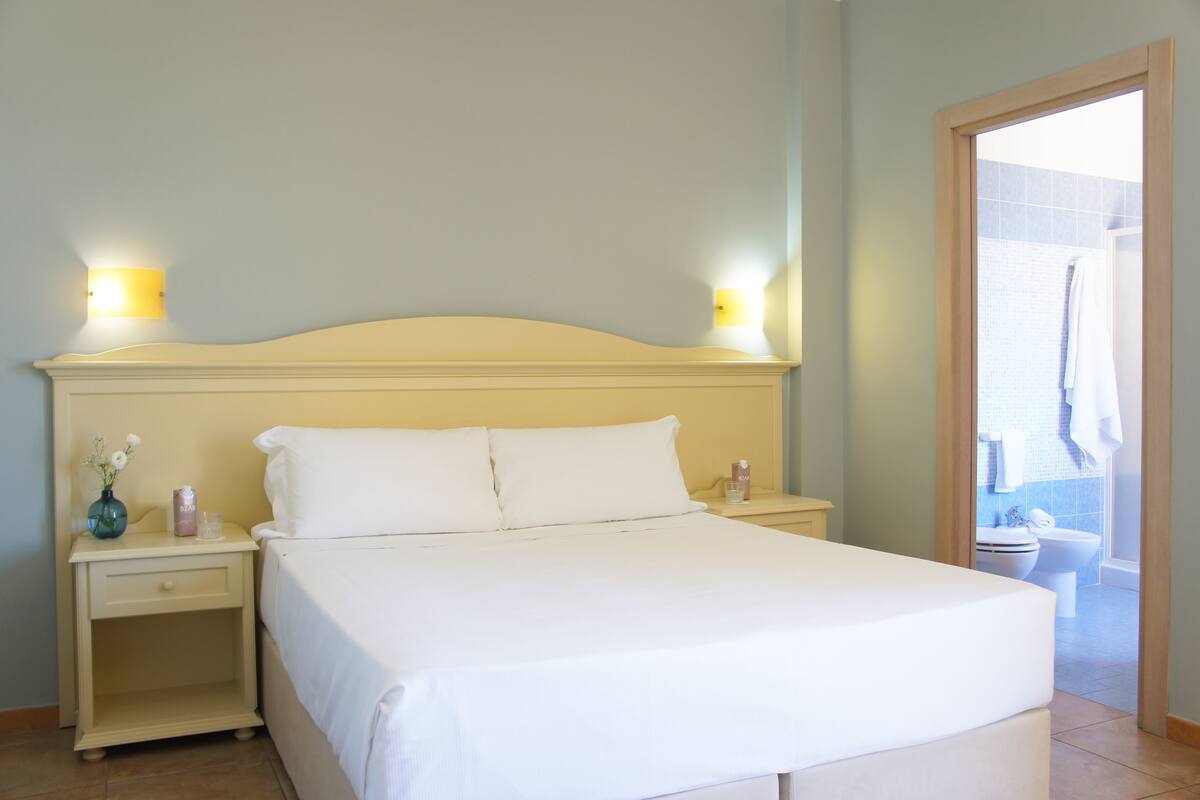 A neatly made bed with white linens, two bedside tables with lamps, and a view of the bathroom through an open door.
