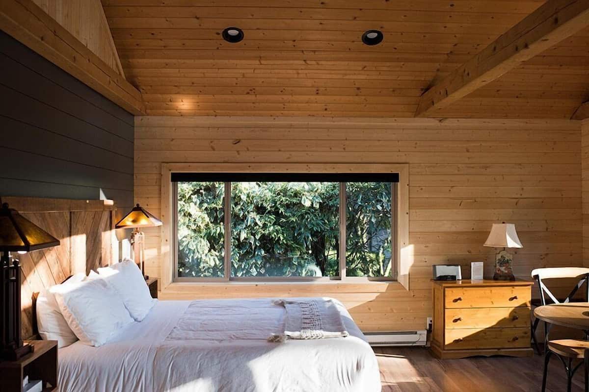 A bedroom with a large window, wooden walls, a bed, bedside lamps, a dresser, and a chair.