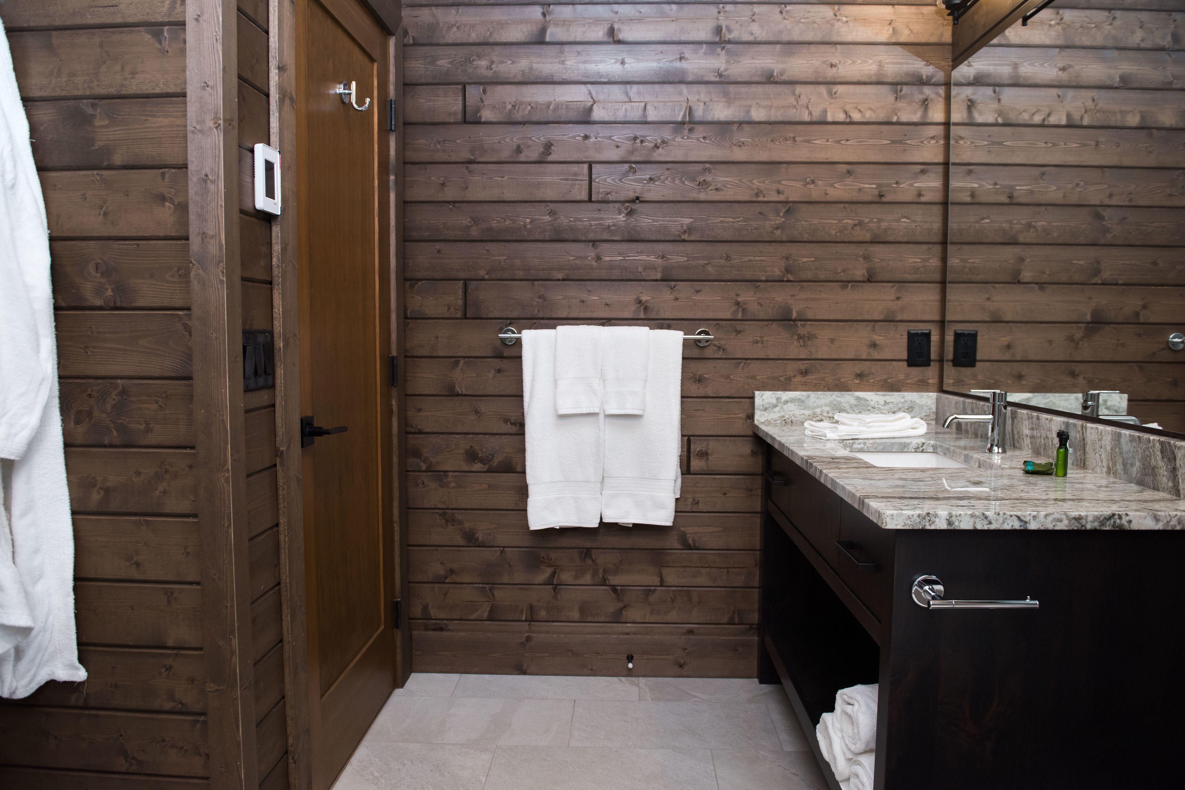 Un baño con paredes y puerta de madera, un perchero con toallas, un lavabo y un espejo.