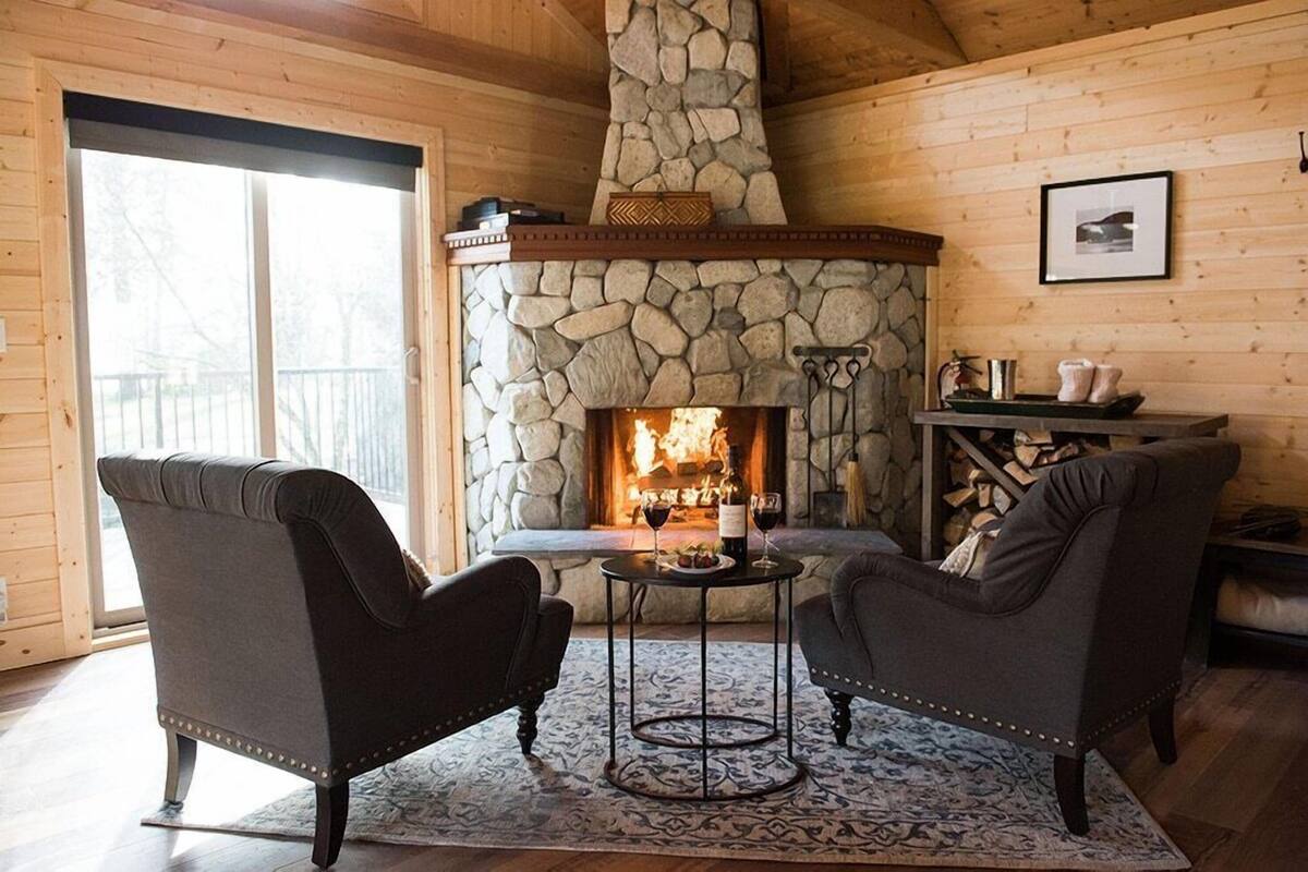 A cozy room with a stone fireplace, two armchairs, a small table with wine glasses, and a view through a large window.