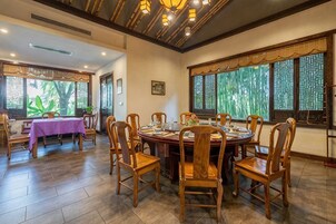 A dining area with a round table and chairs, large windows with decorative screens, and a view of greenery outside.