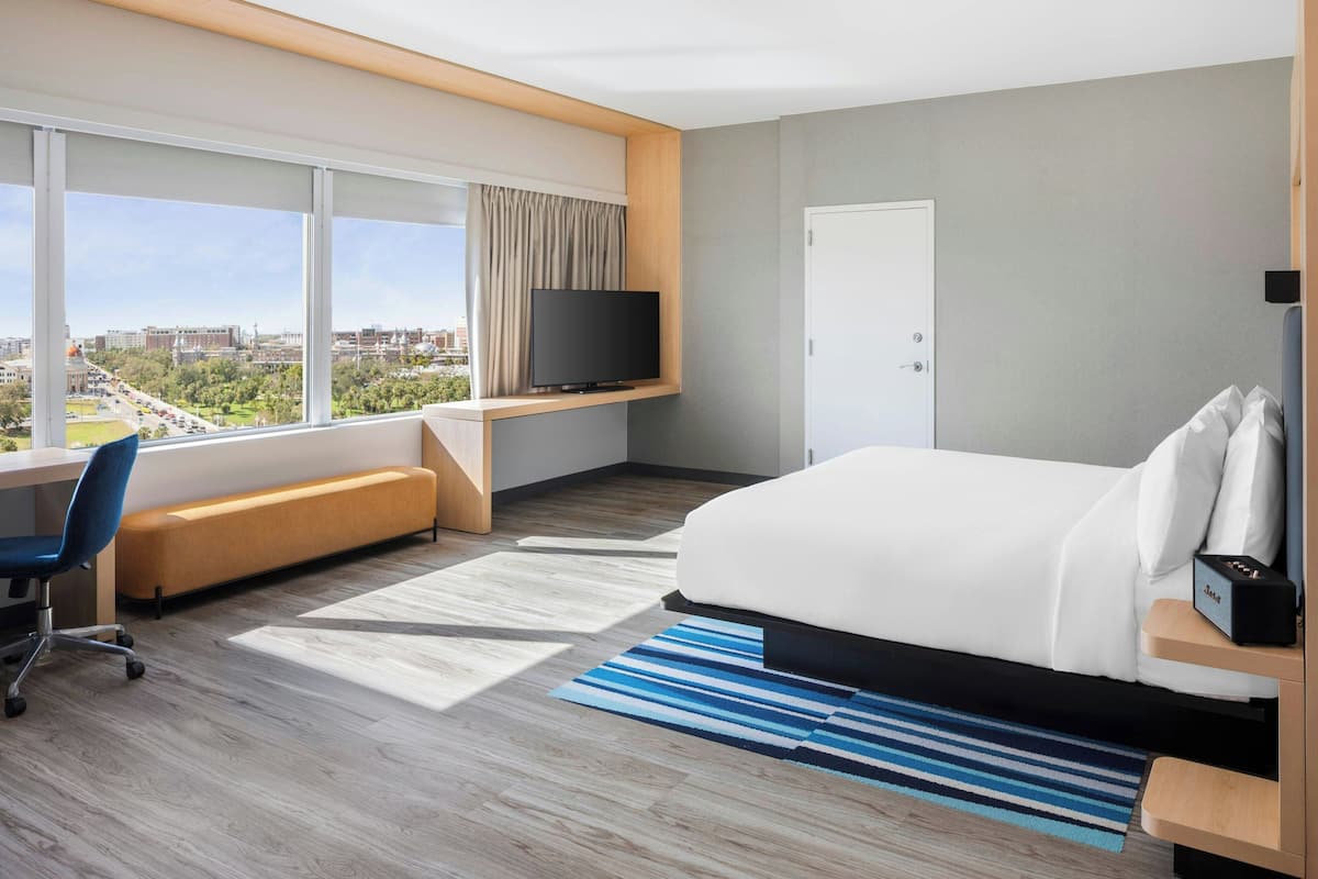 A modern hotel room with a large bed, a desk with a chair, a television, and a view of the cityscape.