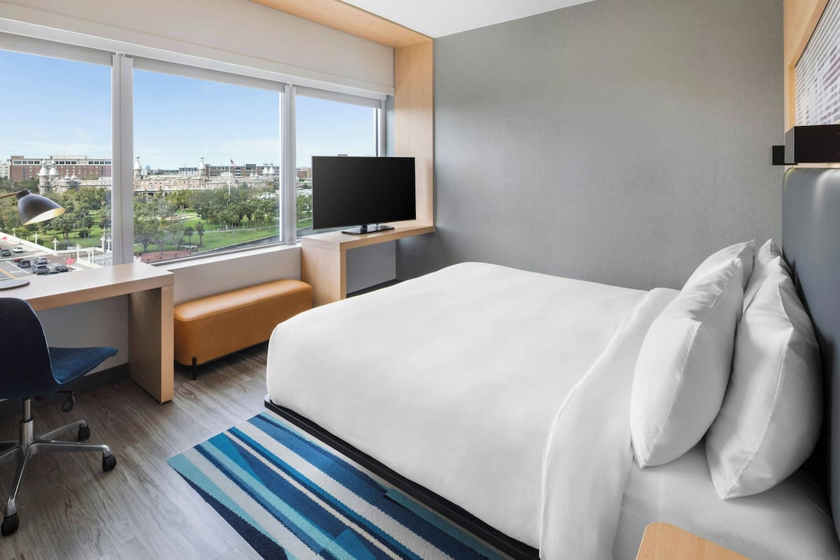 A hotel room with a large bed, a desk with a computer, a TV, and a view of a cityscape.