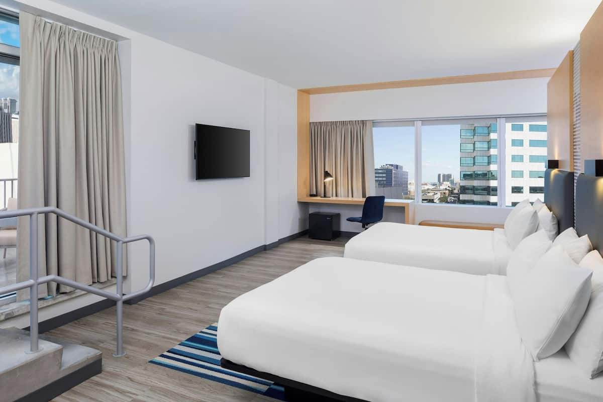 A hotel room with two white beds, a desk, a chair, a TV, and a large window with a city view.
