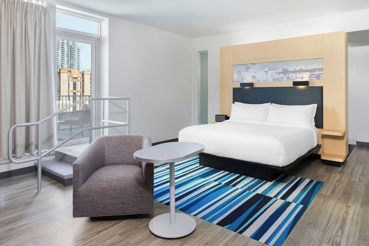 A modern hotel room with a bed, a round table, a chair, and a view of buildings through a window.
