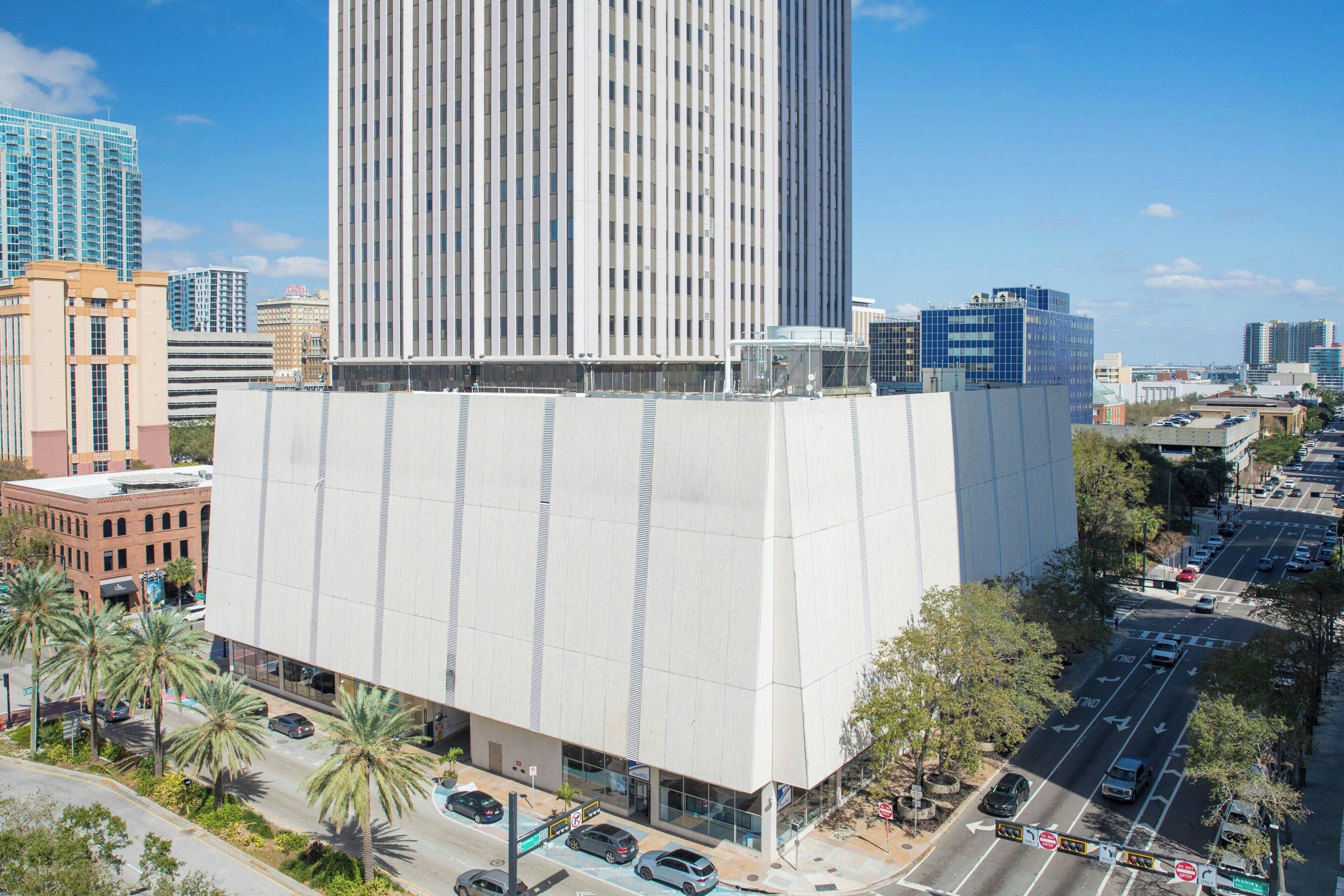 A modern building with a flat facade and large windows, surrounded by palm trees and other buildings.